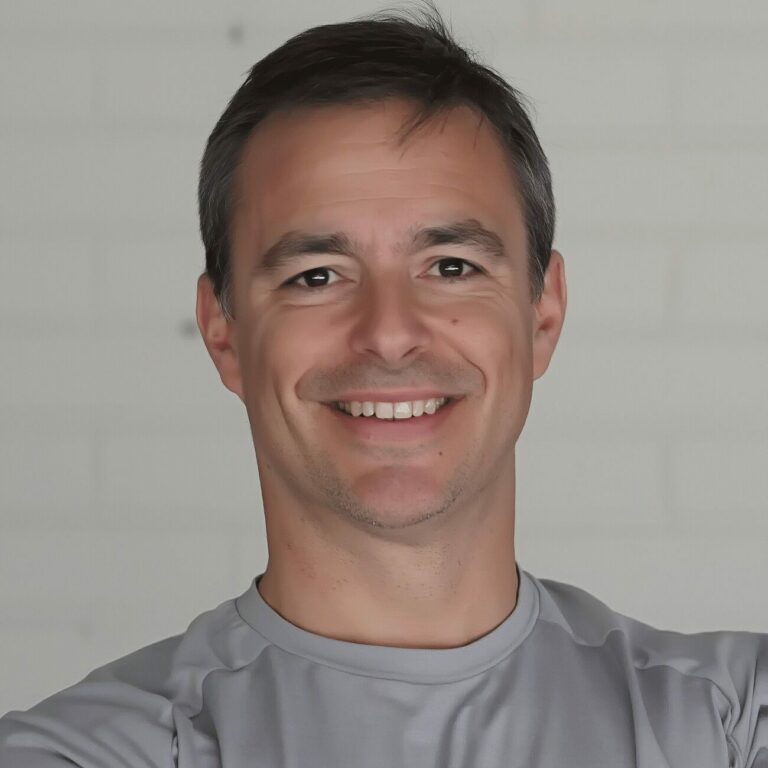 Smiling golf coach in a grey shirt standing confidently with arms crossed, against a light background.