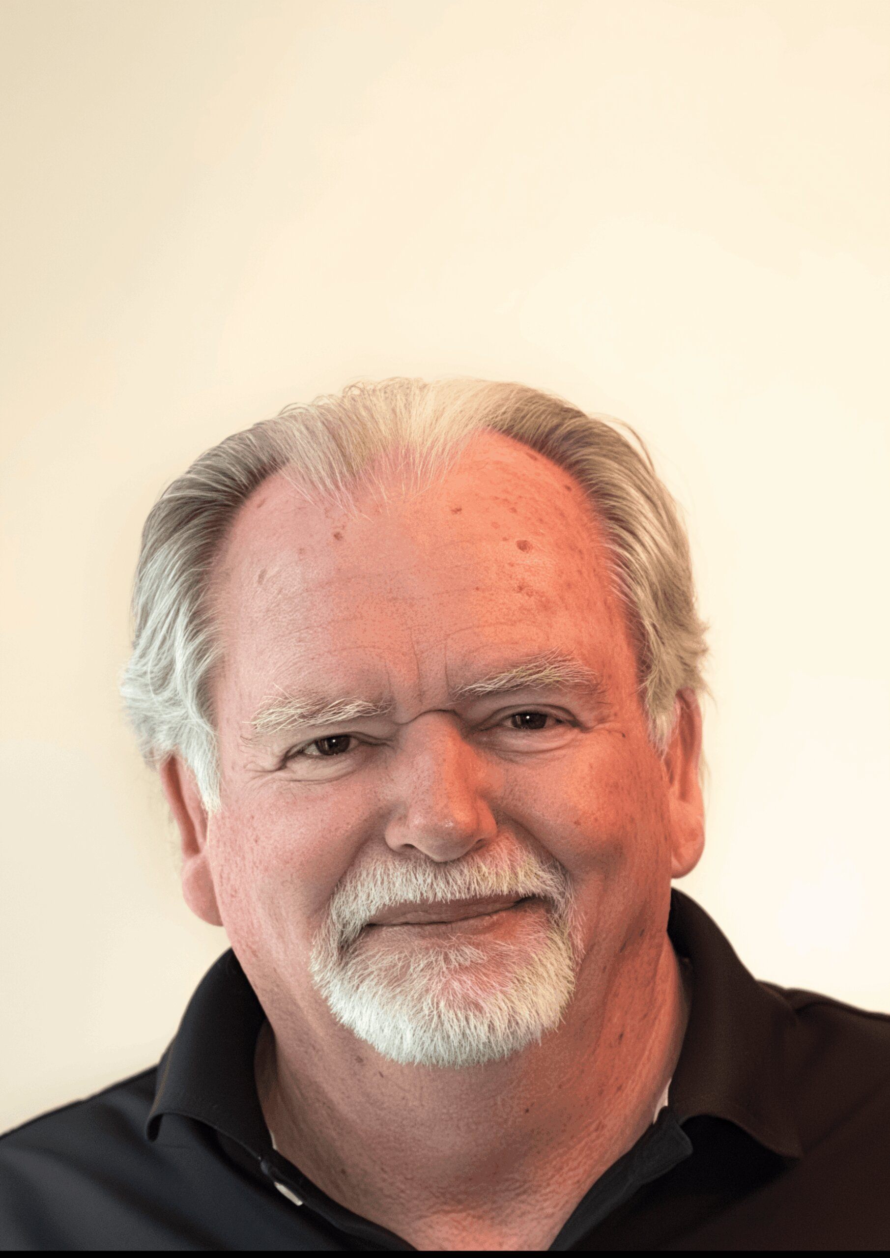 Senior golf mentor with white hair and beard, wearing a black polo shirt, smiling warmly in front of a light background.
