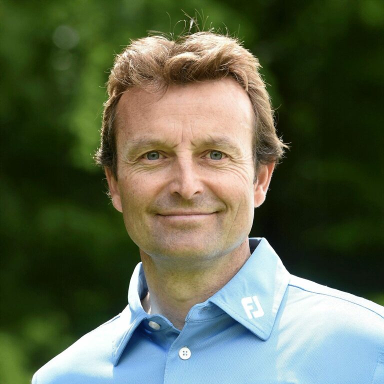 Smiling golf professional at the American Golf Academy’s Junior Golf Academy, wearing a light blue shirt, standing outdoors on a sunny day with a green background.