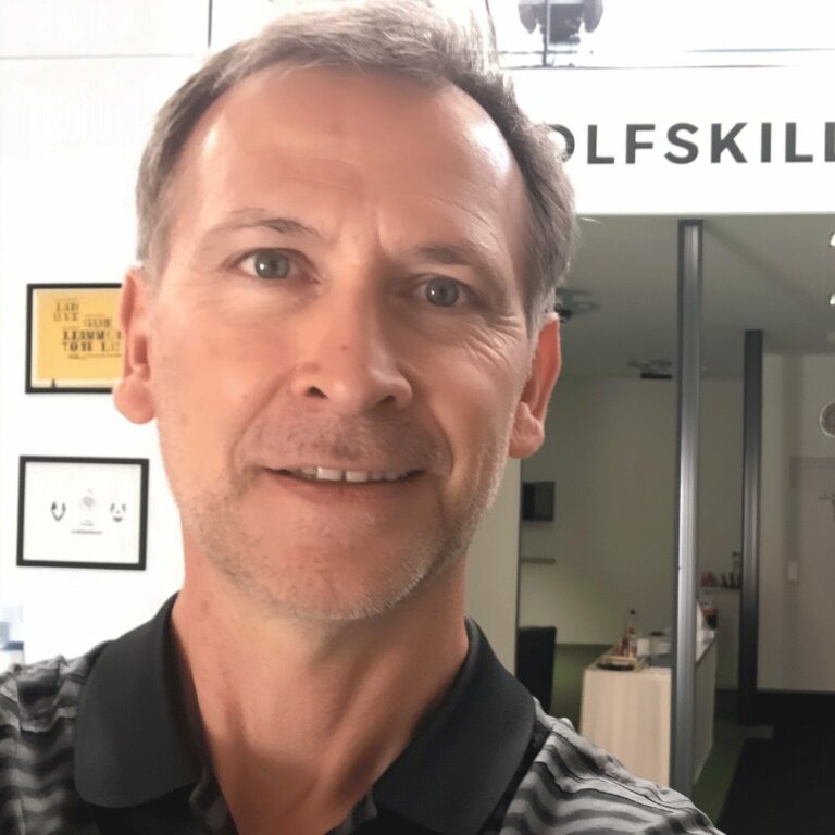 Smiling golf instructor in a striped polo shirt, standing in front of an indoor golf training facility.