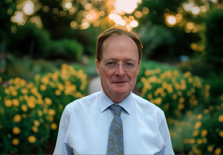 Distinguished gentleman in a light blue shirt and patterned tie, standing in a garden at sunset.